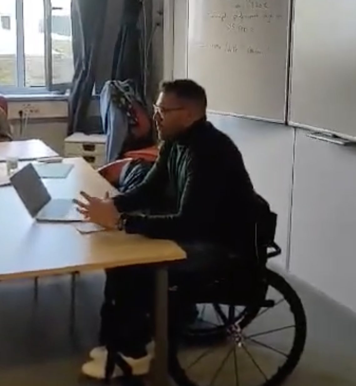 Un homme assis à une table sur un fauteuil roulant, devant un tableau blanc et avec un ordinateur portable devant lui.