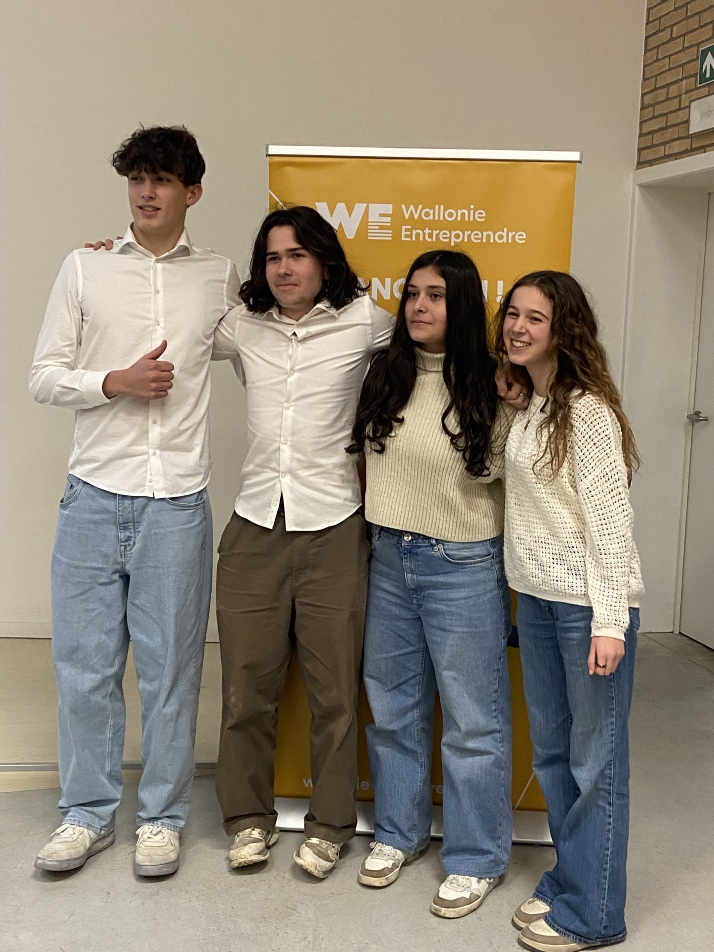 Quatre élèves posent devant un panneau roll-up de "Wallonie Entreprendre". Celui à gauche de la photo a son pouce levé.