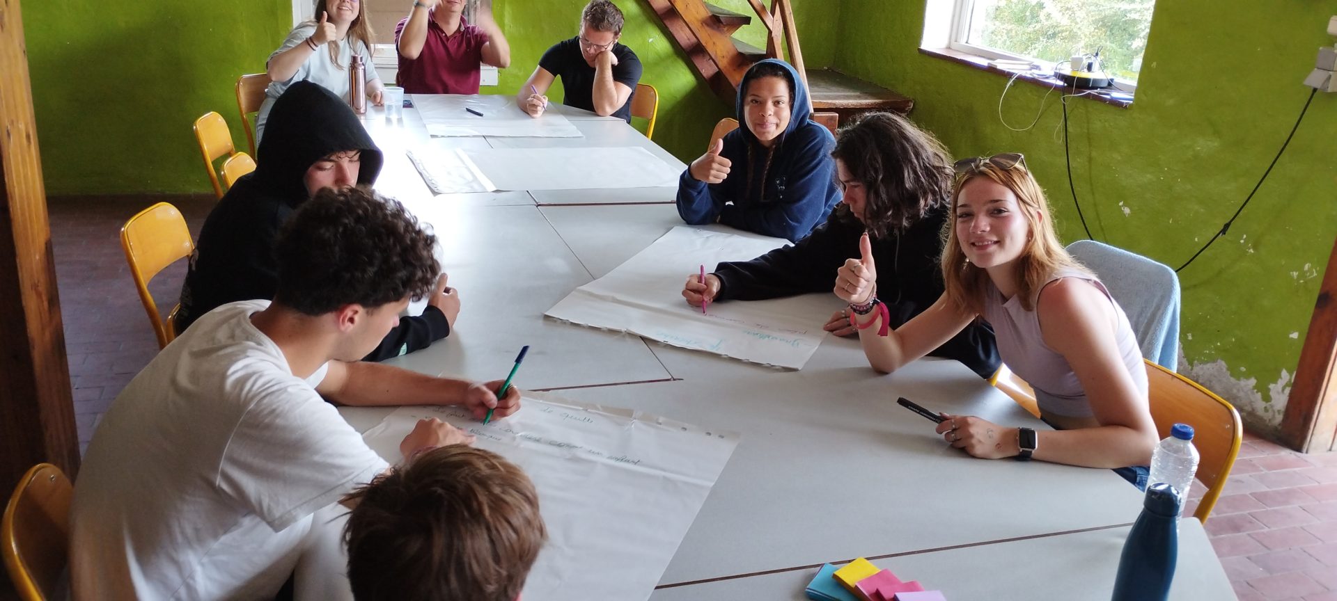 Plusieurs élèves sont réunis autour d'une table dans une salle aux murs verts. Ils travaillent ensemble en écrivant sur des grandes feuilles blanches. Plusieurs d'entre eux regardent l'appareil photo et lèvent leur pouce.
